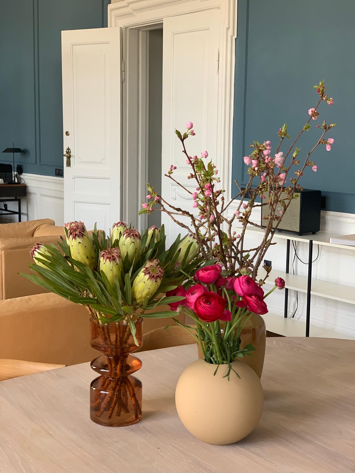 Dekorationer med proteaer, ranunkler og blomstergrene på et bord i et lokale med blå paneler