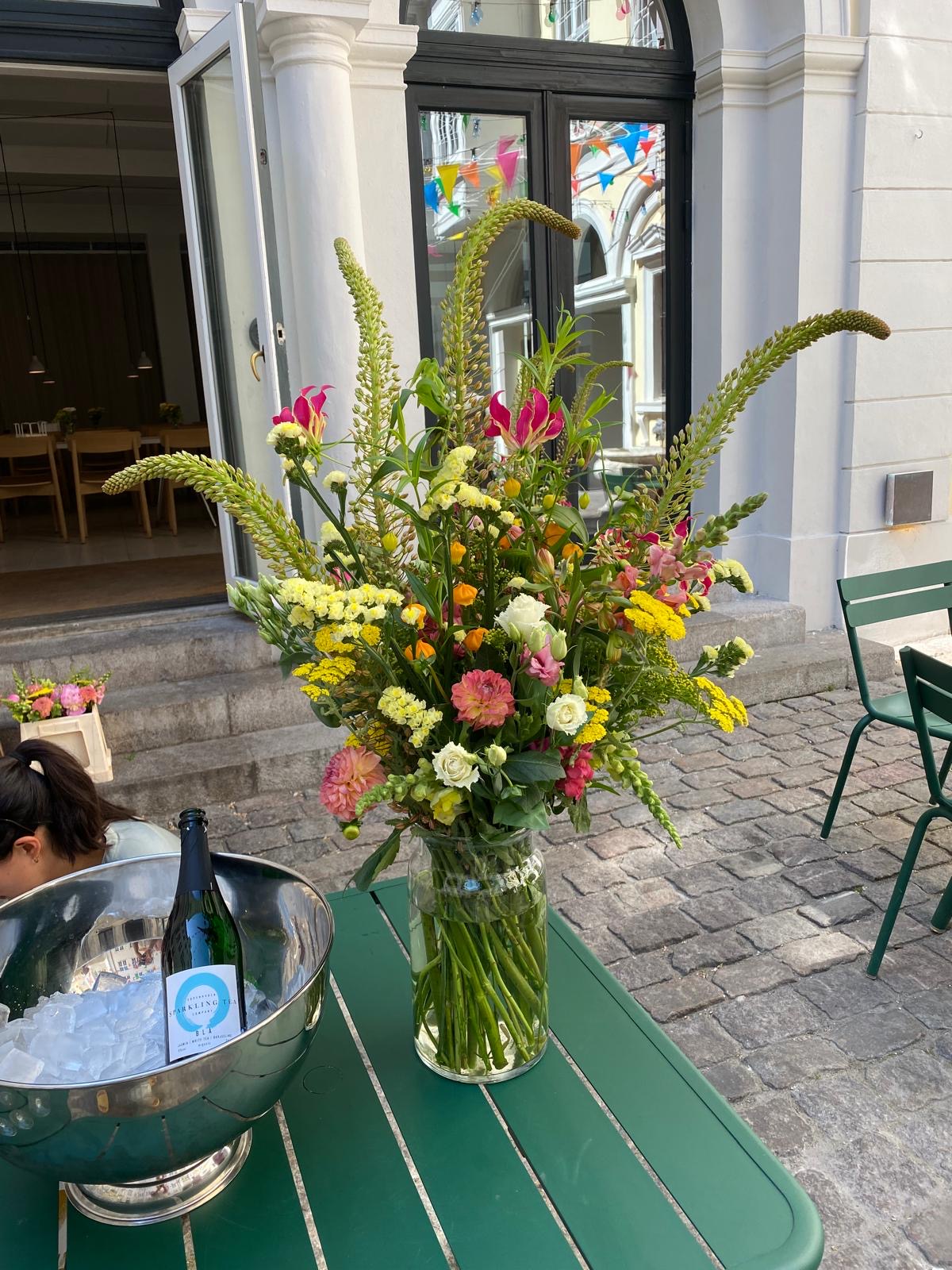 Stor blomsterdekoration i glasvase på udeservering med grønne stole
