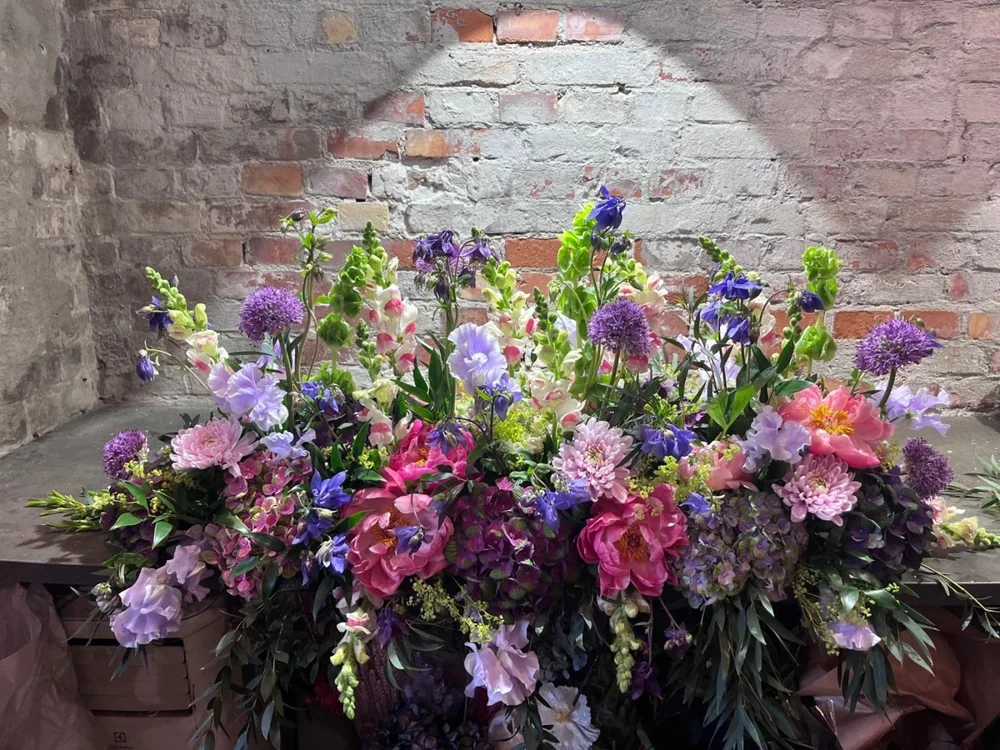 Frodigt arrangement med lyserøde og lilla blomster foran en murstensvæg