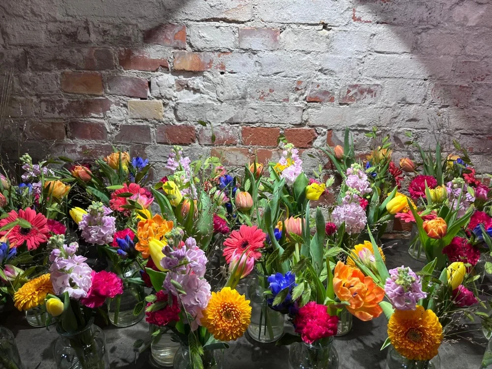 Farverige buketter med solsikker, roser og lilla blomster foran en murstensvæg