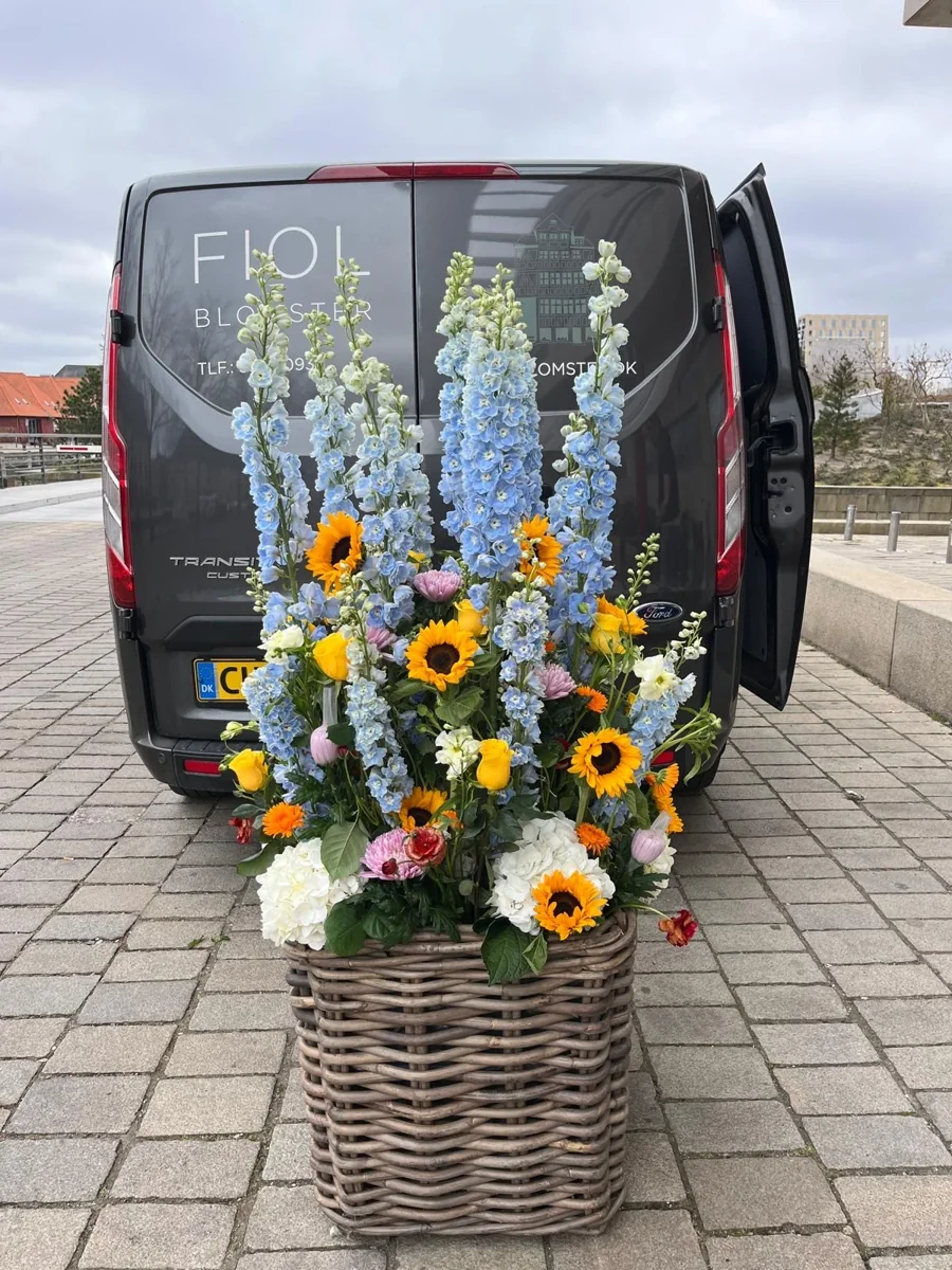 Højt arrangement med solsikker, blå ridderspore og hvide blomster ved Fiols varevogn